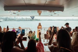 Corporate event hosted on AO Cruises yacht with guests applauding a speaker on deck, overlooking Sydney Harbour and city skyline.